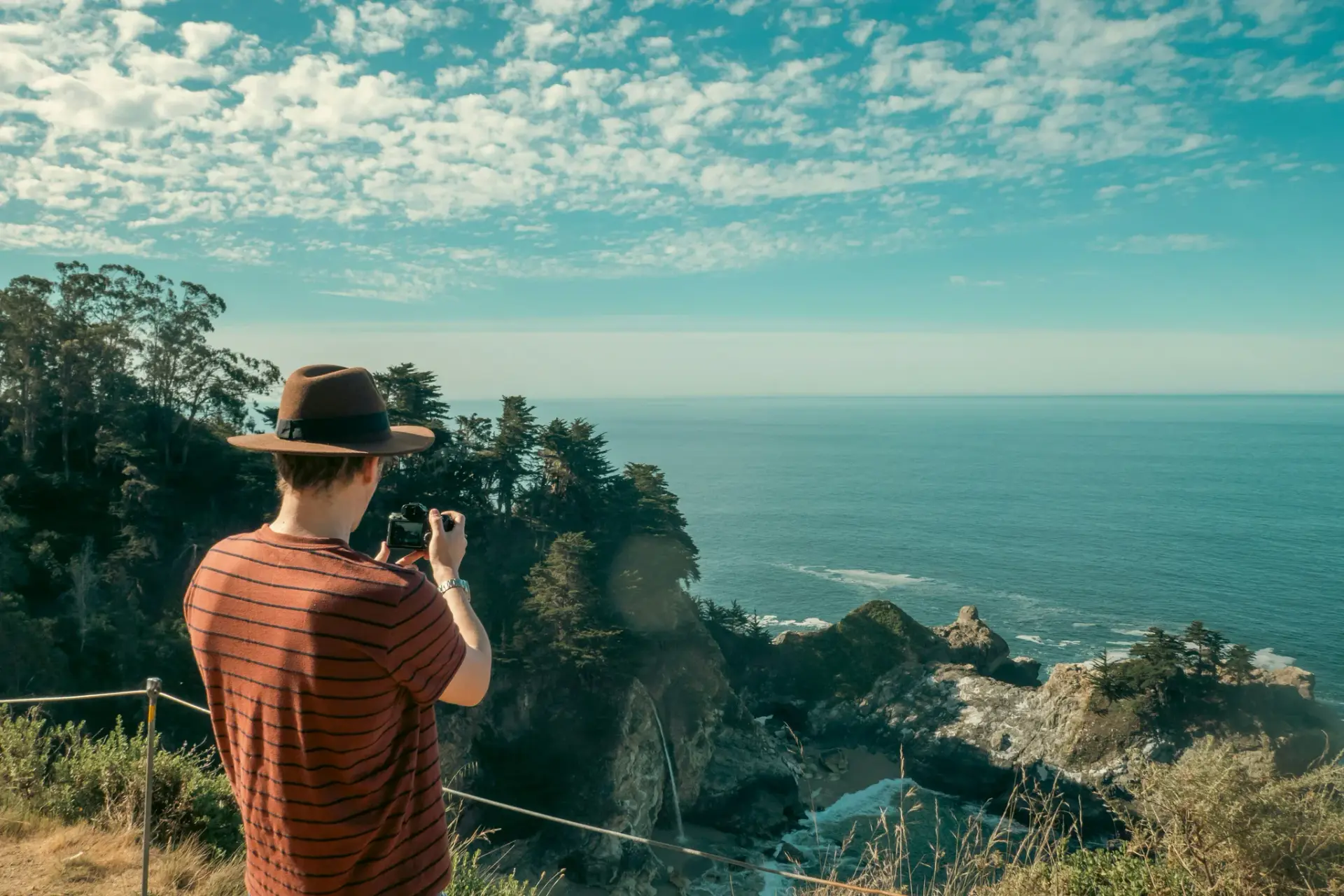 Man in hat photographs ocean view with smartphone.